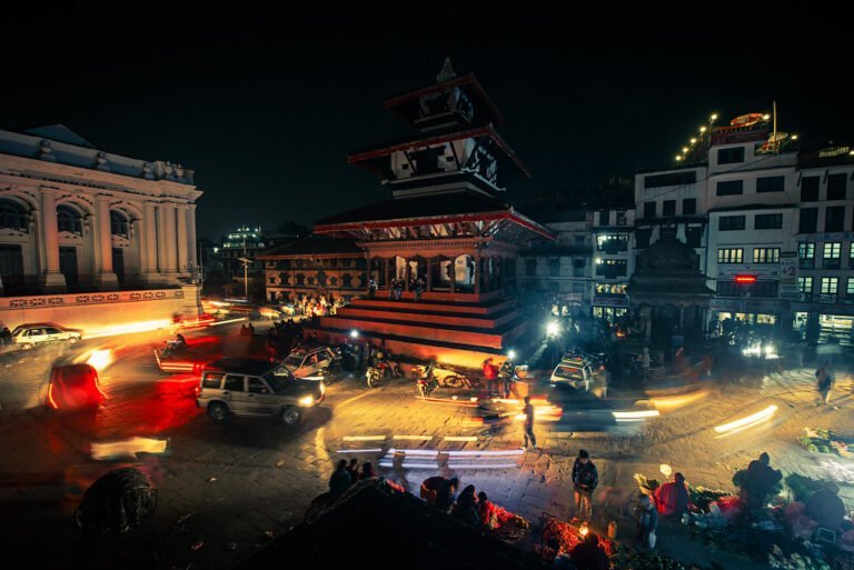 A bustling night scene at a city square with a traditional multi-tiered temple lit by ambient light, surrounded by motion-blurred vehicles and pedestrians, with some people gathered around vendors on the street.