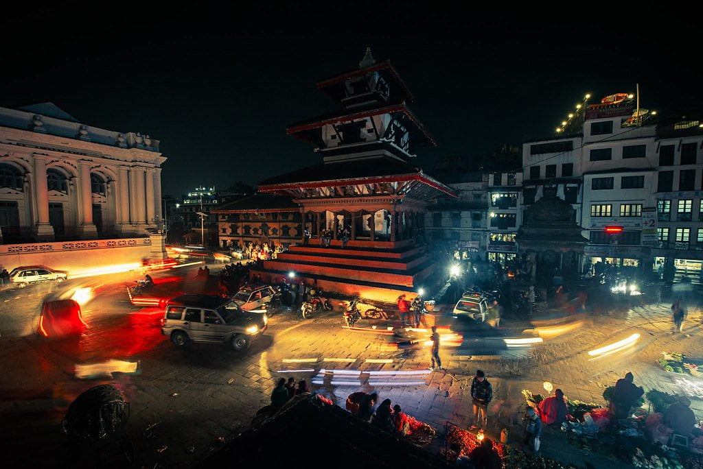 A bustling night scene at a city square with a traditional multi-tiered temple lit by ambient light, surrounded by motion-blurred vehicles and pedestrians, with some people gathered around vendors on the street.