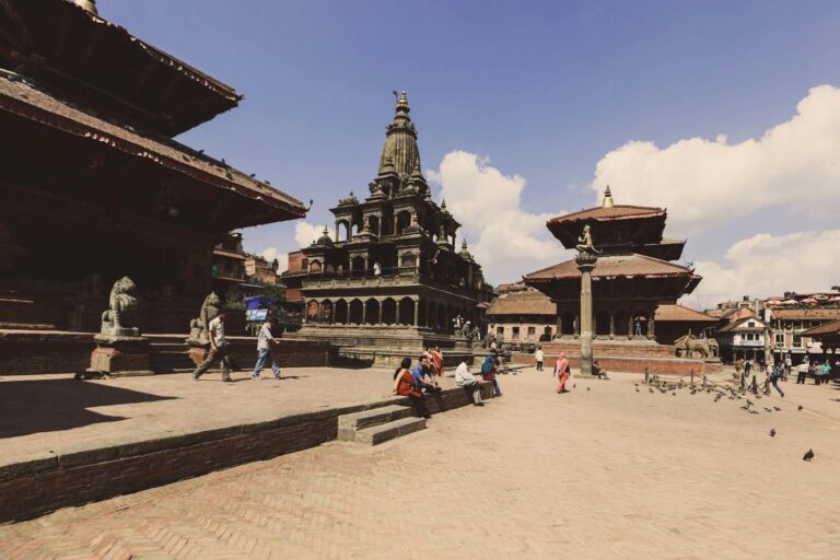 Ancient temples and structures in Durbar Square with people walking and sitting around on a sunny day, clear blue sky above, and pigeons scattered on the ground in Kathmandu, Nepal.
