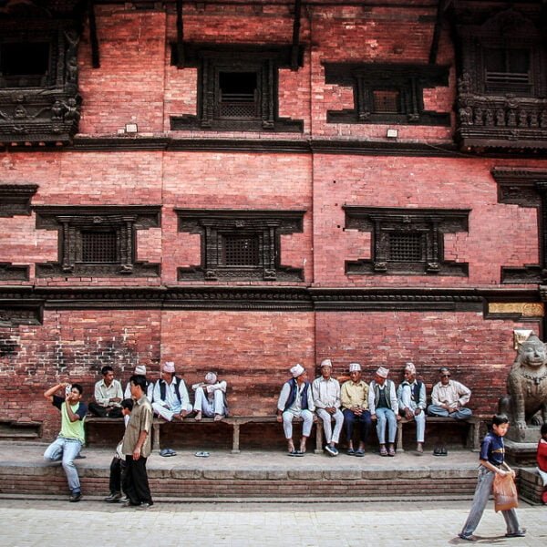 A group of people sitting and standing in front of a traditional Nepalese building with intricate wood carvings, while a bird flies by and pedestrians walk in the foreground.