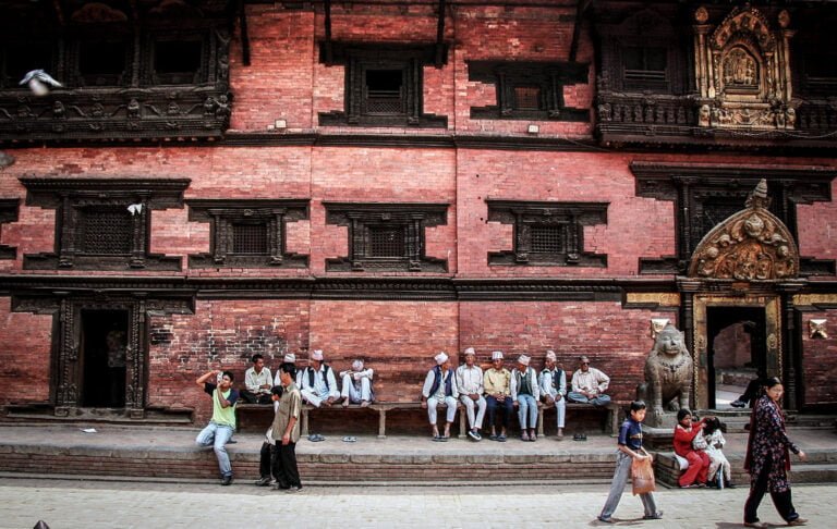 A group of people sitting and standing in front of a traditional Nepalese building with intricate wood carvings, while a bird flies by and pedestrians walk in the foreground.