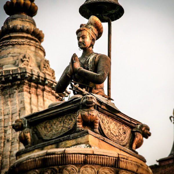 Statue of a historical figure with hands pressed together in a gesture of respect, standing atop an ornate pedestal with traditional Nepalese architectural elements and smaller spires in the background.