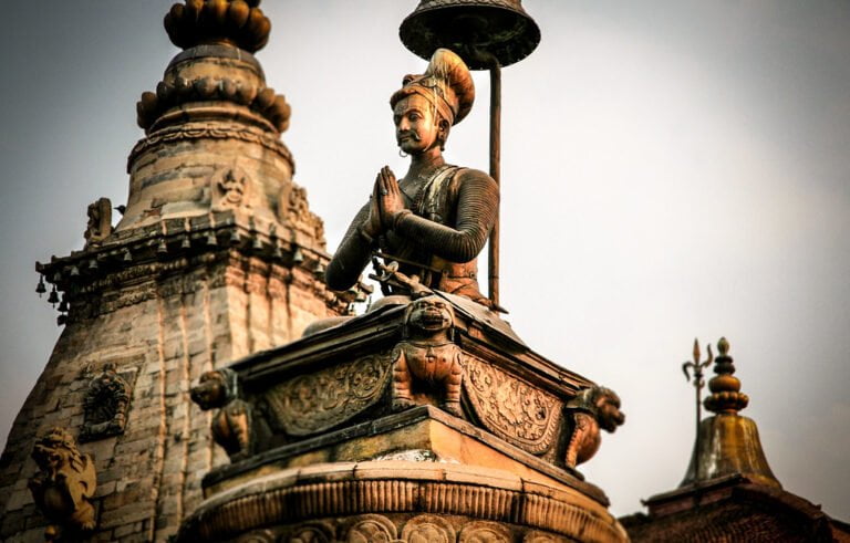 Statue of a historical figure with hands pressed together in a gesture of respect, standing atop an ornate pedestal with traditional Nepalese architectural elements and smaller spires in the background.