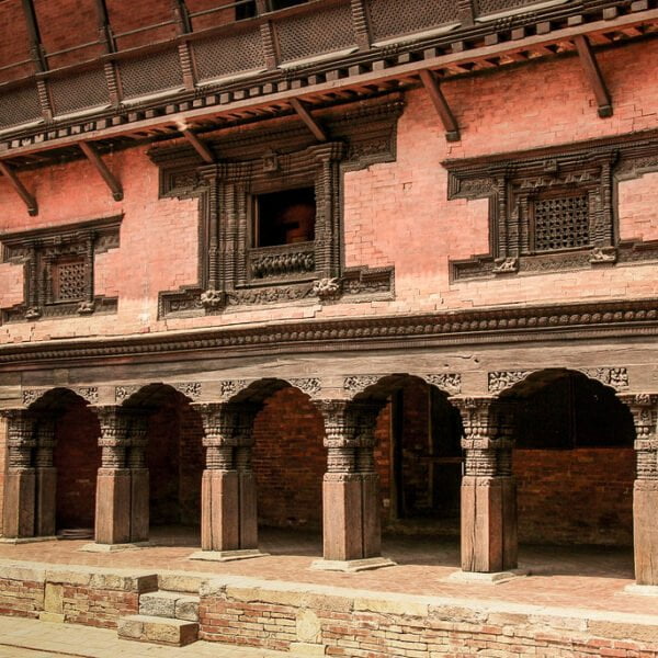 Alt text: An ancient brick building with intricate wooden carvings on window frames and columns, featuring a row of arches along the ground floor, characteristic of traditional Nepalese architecture.