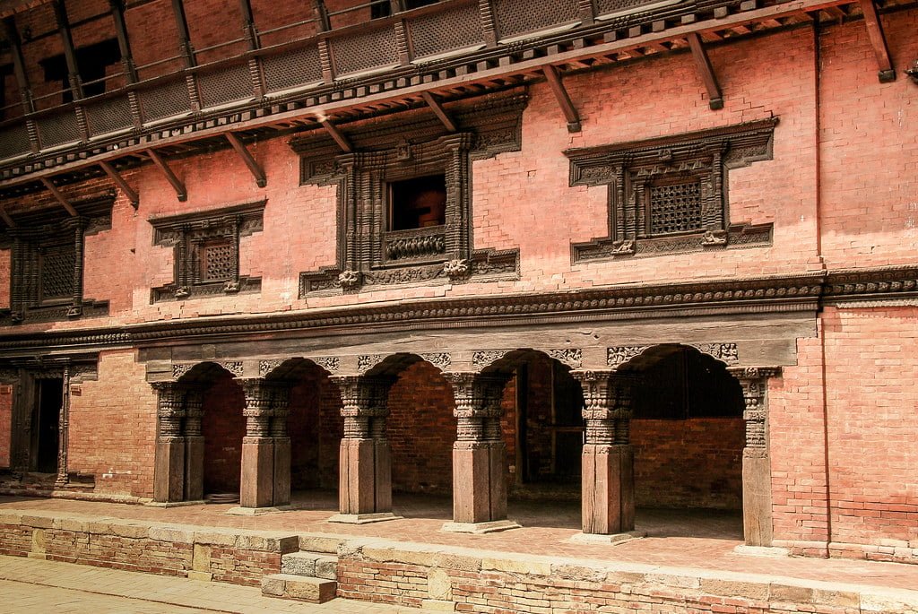 Alt text: An ancient brick building with intricate wooden carvings on window frames and columns, featuring a row of arches along the ground floor, characteristic of traditional Nepalese architecture.