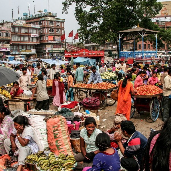A bustling outdoor market with numerous people trading fruits and vegetables in a crowded urban setting, with umbrellas providing shade and a backdrop of multistory buildings displaying signs and banners.