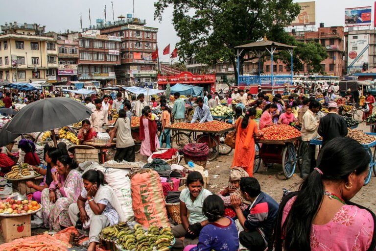 A bustling outdoor market with numerous people trading fruits and vegetables in a crowded urban setting, with umbrellas providing shade and a backdrop of multistory buildings displaying signs and banners.
