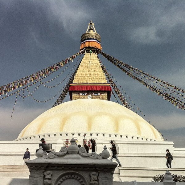 Boudhanath Stupa in Kathmandu, Nepal, with colorful prayer flags radiating from the top under a cloudy sky, while visitors explore the surroundings.