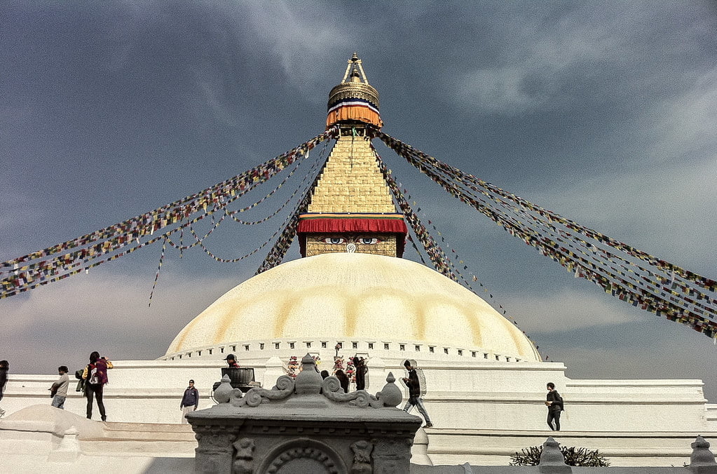 Boudhanath Stupa in Kathmandu, Nepal, with colorful prayer flags radiating from the top under a cloudy sky, while visitors explore the surroundings.