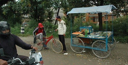 Two street vendors with carts alongside a road, one cart covered with a blue tarp, as a motorcyclist passes by in the foreground.