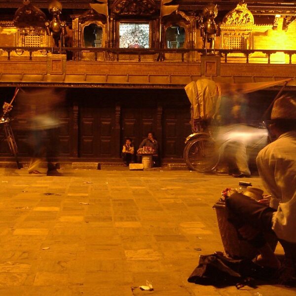 A night scene in a traditional square with motion blur of passing individuals, featuring a seated person in the foreground, ornate wooden architecture in the background, and ambient warm lighting.