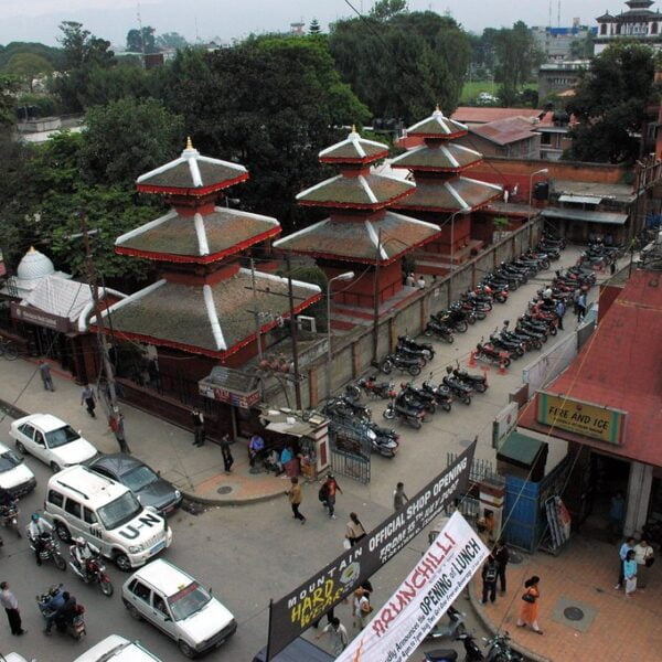 A busy street scene with traditional Nepalese architecture, featuring multi-tiered pagoda-style temples and a line of parked motorcycles beside a red building with "FIRE AND ICE" sign, with cars and pedestrians scattered throughout.