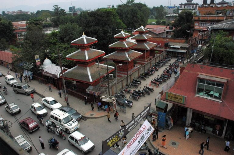 A busy street scene with traditional Nepalese architecture, featuring multi-tiered pagoda-style temples and a line of parked motorcycles beside a red building with "FIRE AND ICE" sign, with cars and pedestrians scattered throughout.
