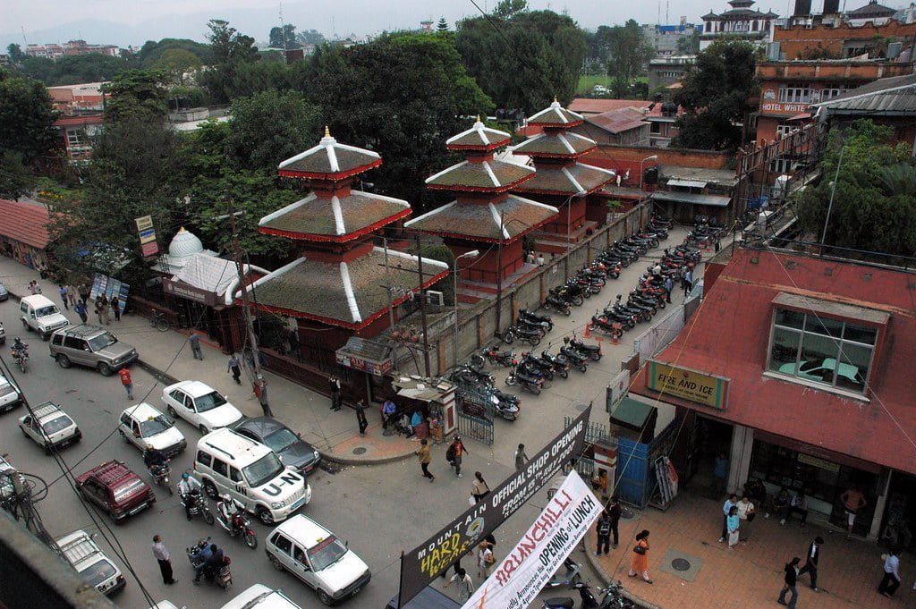 A busy street scene with traditional Nepalese architecture, featuring multi-tiered pagoda-style temples and a line of parked motorcycles beside a red building with "FIRE AND ICE" sign, with cars and pedestrians scattered throughout.
