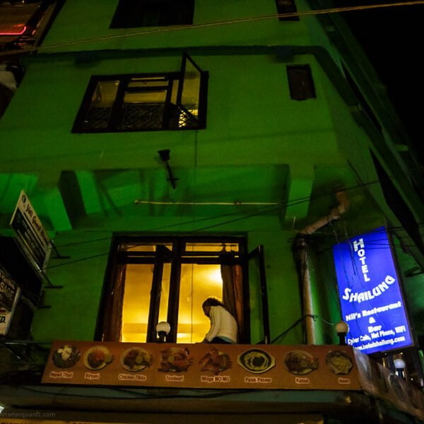 A person is visible through the first-floor window of a green-lit building at night, with various signs for guest homes, treks and expeditions, and a hotel in Kathmandu.