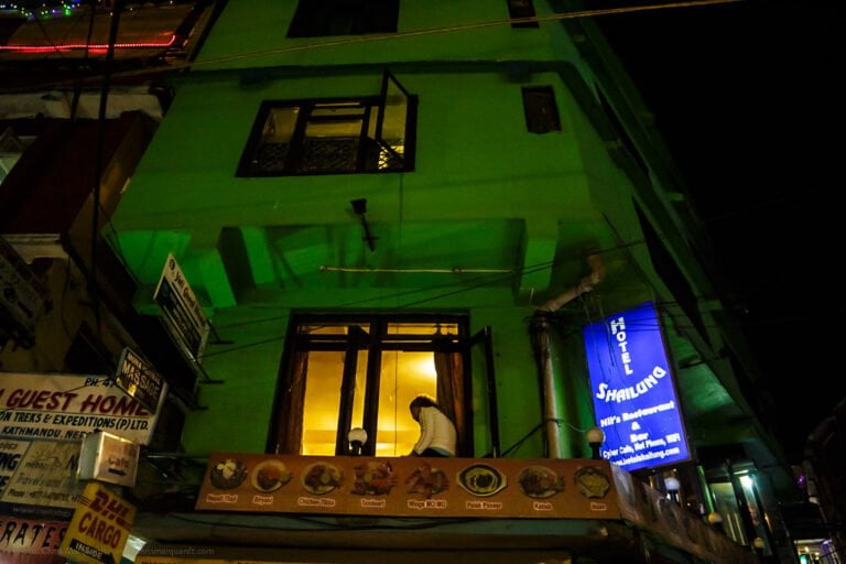 A person is visible through the first-floor window of a green-lit building at night, with various signs for guest homes, treks and expeditions, and a hotel in Kathmandu.
