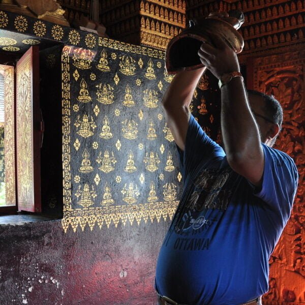 A man in a blue shirt is pouring water from a clay pot above his head inside a room with traditional golden wall patterns and a carved wooden door frame.
