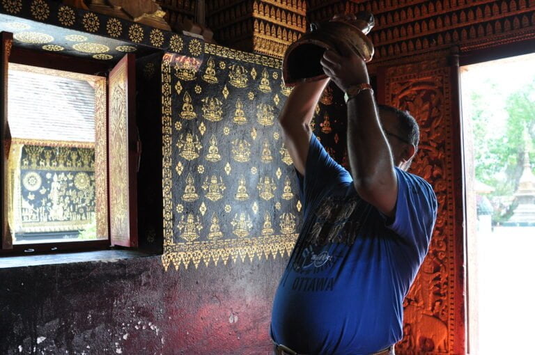 A man in a blue shirt is pouring water from a clay pot above his head inside a room with traditional golden wall patterns and a carved wooden door frame.