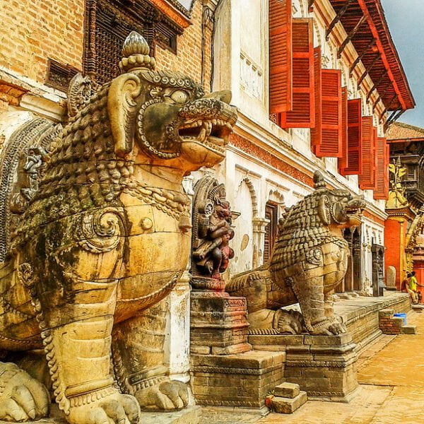 Stone sculptures of mythological creatures guard an entrance in a historic square with red-shuttered buildings and visitors walking in the background.