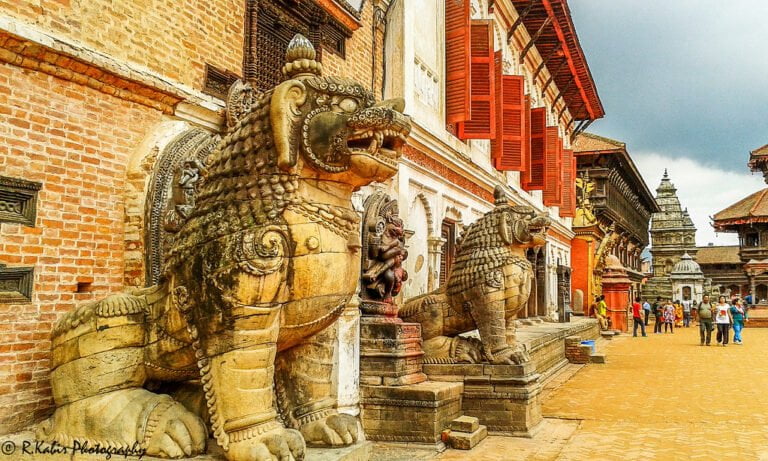 Stone sculptures of mythological creatures guard an entrance in a historic square with red-shuttered buildings and visitors walking in the background.