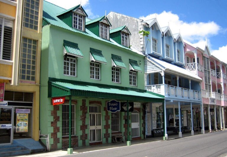 A colorful street lined with two-story Caribbean-style buildings with shops on the ground floor and residential spaces on the top, under a bright blue sky.