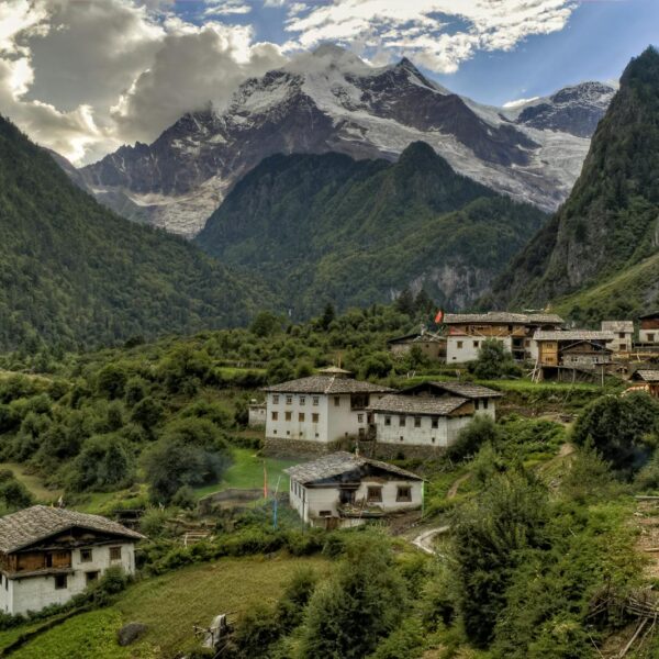 A mountainous landscape with a rural village consisting of traditional houses, surrounded by lush greenery and a backdrop of snow-capped peaks and glaciers under a cloudy sky.