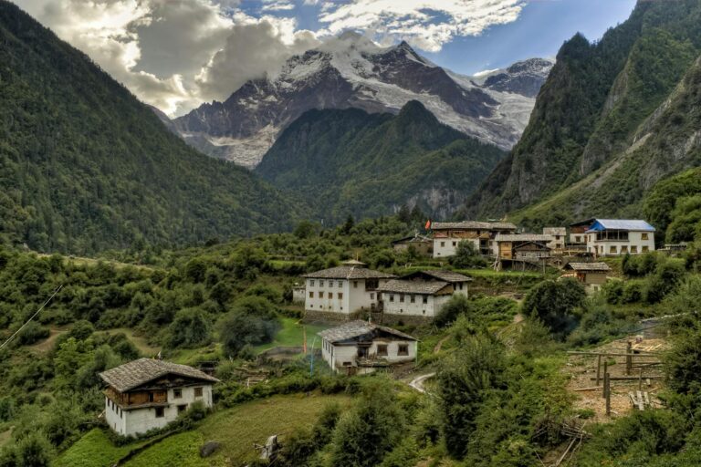A mountainous landscape with a rural village consisting of traditional houses, surrounded by lush greenery and a backdrop of snow-capped peaks and glaciers under a cloudy sky.
