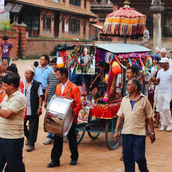 A cultural procession with musicians playing trumpets and drums, followed by a crowd of people, with a decorated carriage in the center, taking place in a traditional square with historic architecture in the background.