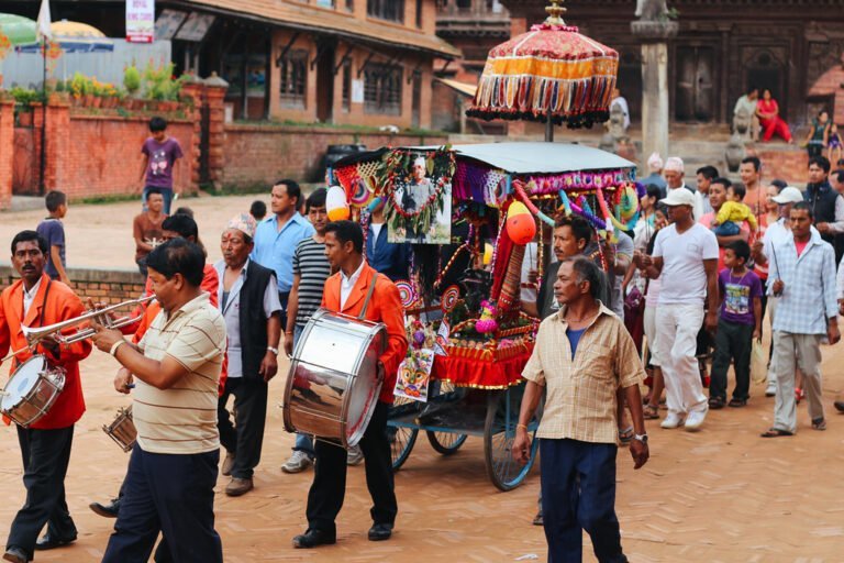 A cultural procession with musicians playing trumpets and drums, followed by a crowd of people, with a decorated carriage in the center, taking place in a traditional square with historic architecture in the background.