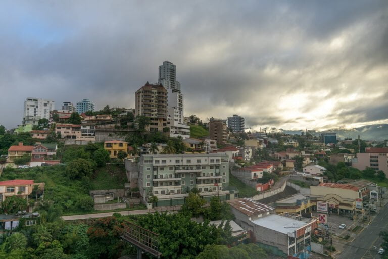 A cityscape with a mix of residential buildings on a hillside and commercial buildings in the foreground, under a cloudy sky with rays of sunlight breaking through.