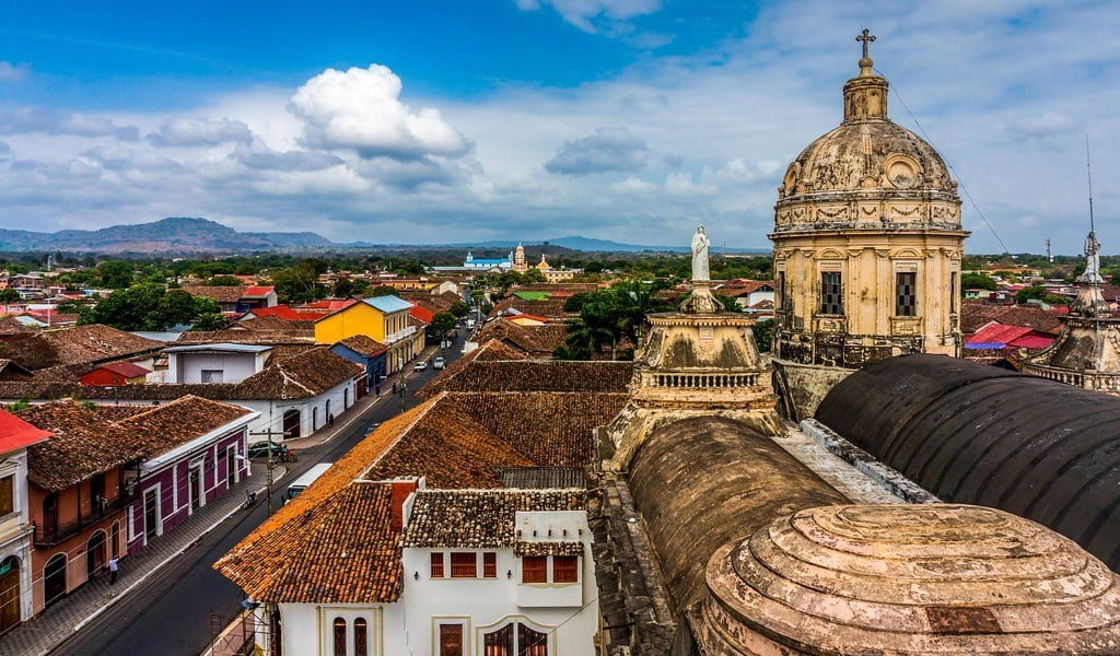 Aerial view of a historic colonial town with red-tiled rooftops, a prominent weathered dome of a cathedral on the right, and mountains in the background under a partly cloudy sky.