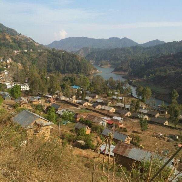 A panoramic view of a rural village nestled in a hilly landscape with a meandering river, terraced fields, and houses with various types of roofing materials.