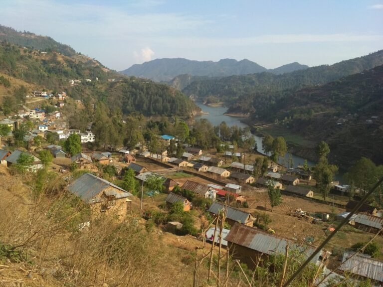 A panoramic view of a rural village nestled in a hilly landscape with a meandering river, terraced fields, and houses with various types of roofing materials.