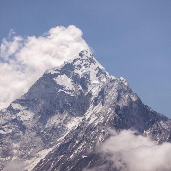 A majestic snow-capped mountain peak piercing through white clouds against a clear blue sky.