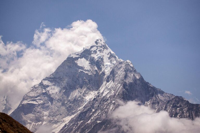 A majestic snow-capped mountain peak piercing through white clouds against a clear blue sky.