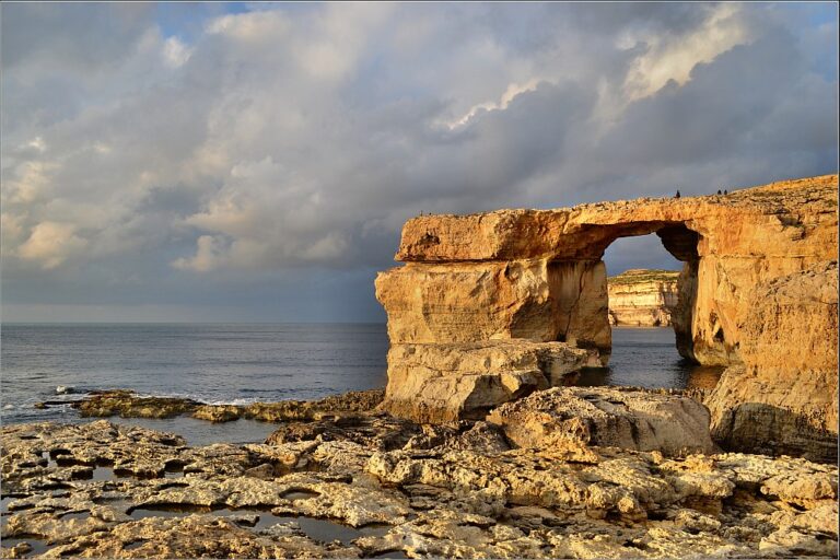 A natural limestone arch on a rocky coastline illuminated by warm sunlight against a backdrop of cloudy skies.