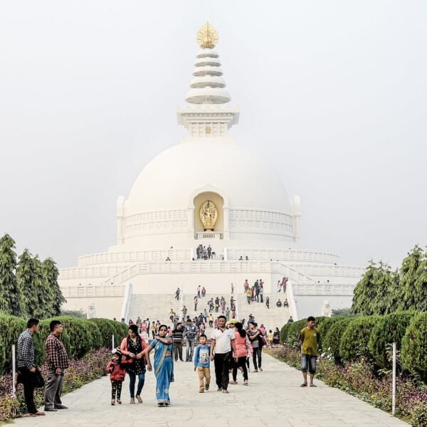 A white stupa with a golden structure on top surrounded by visitors on pathways lined with shrubs and trees under a hazy sky.