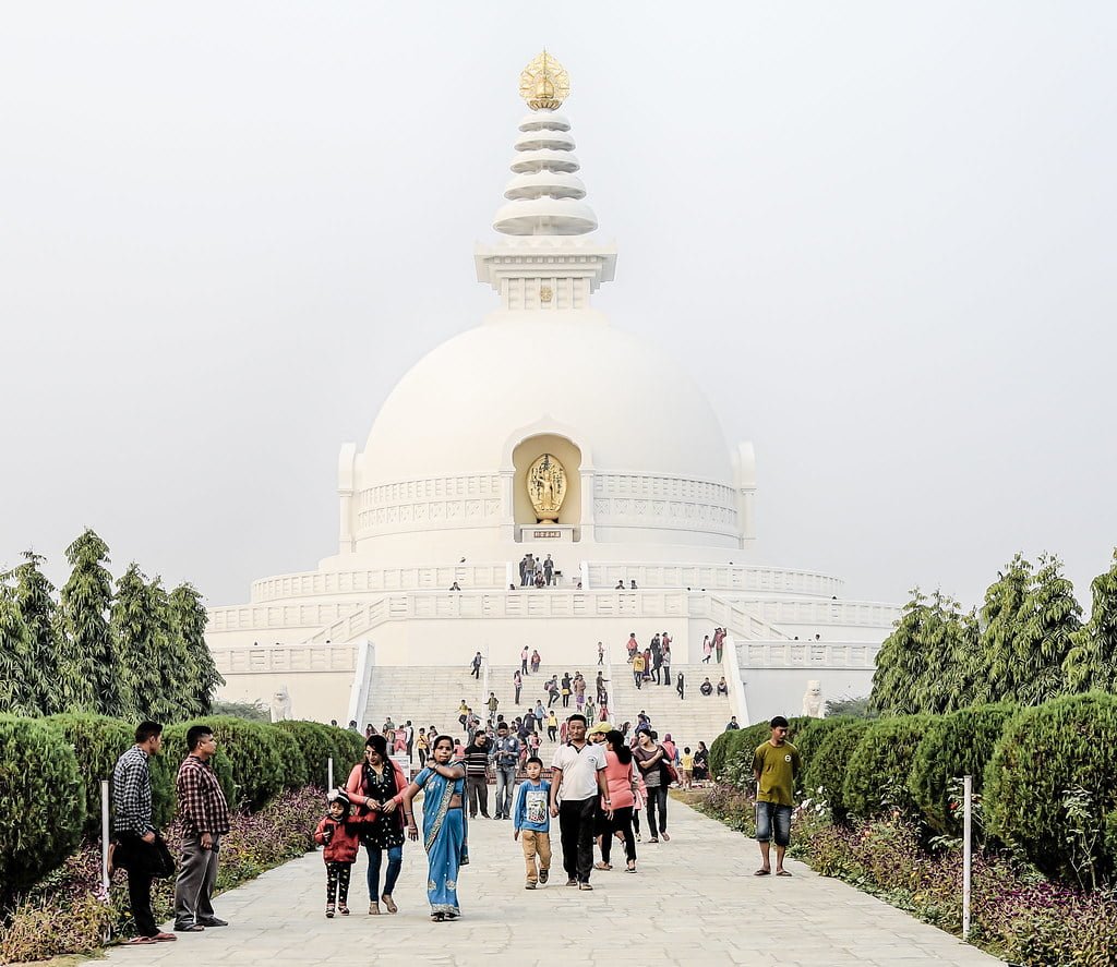 A white stupa with a golden structure on top surrounded by visitors on pathways lined with shrubs and trees under a hazy sky.