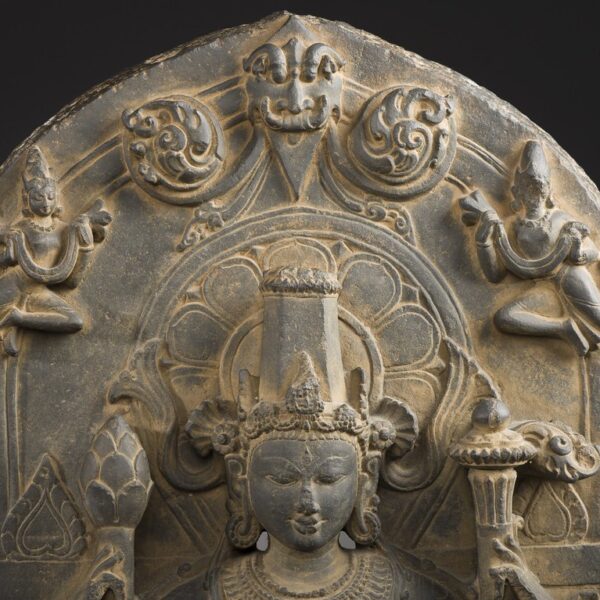 An intricately carved stone relief depicting a central figure with a serene expression, flanked by two smaller figures in dance-like postures, all set within an arched framework with floral and geometric designs.