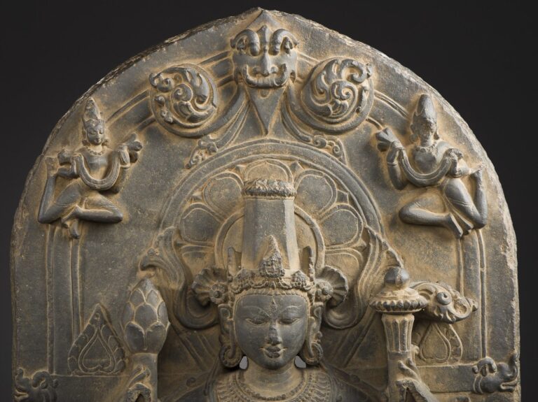 An intricately carved stone relief depicting a central figure with a serene expression, flanked by two smaller figures in dance-like postures, all set within an arched framework with floral and geometric designs.