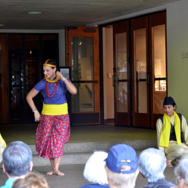 Four performers in traditional Southeast Asian attire are dancing and posing in front of an audience seated on the ground, with a building entrance and a sign reading "BERKELEY CENTER GALLERY" in the background.