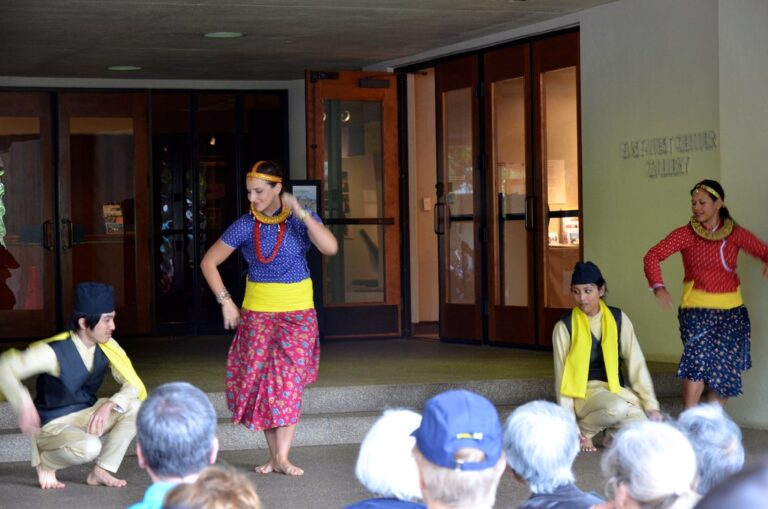 Four performers in traditional Southeast Asian attire are dancing and posing in front of an audience seated on the ground, with a building entrance and a sign reading "BERKELEY CENTER GALLERY" in the background.