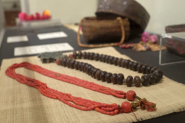 Traditional necklaces displayed on a burlap surface with cultural artifacts blurred in the background.
