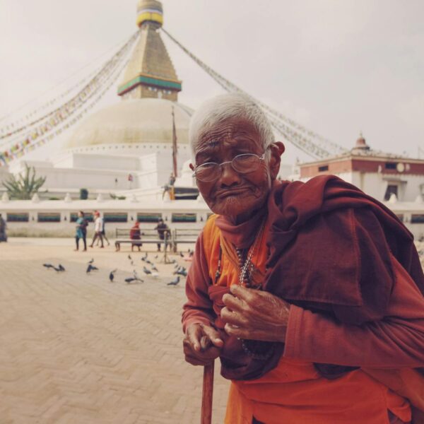 An elderly person in traditional orange and maroon robes with glasses standing in front of the Boudhanath Stupa in Kathmandu, Nepal, with pigeons and people in the background.