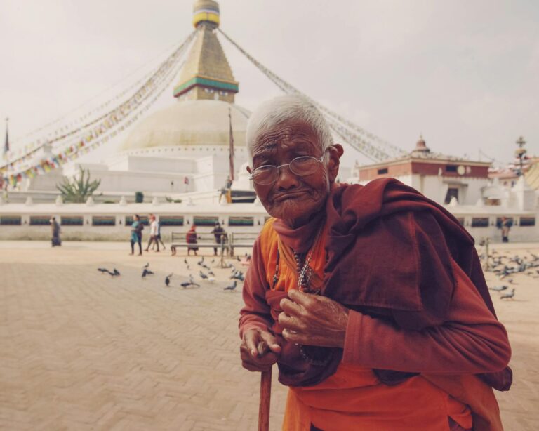 An elderly person in traditional orange and maroon robes with glasses standing in front of the Boudhanath Stupa in Kathmandu, Nepal, with pigeons and people in the background.