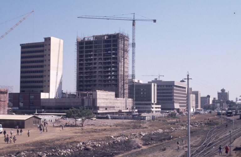 Urban construction site with multiple buildings, a tall crane, people walking nearby, and railroad tracks in the foreground.