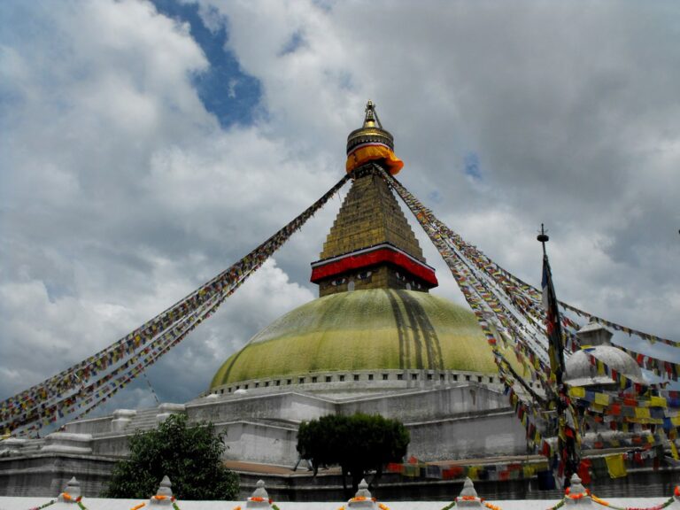 Boudhanath Stupa in Nepal with colorful prayer flags extending from the spire under a cloudy sky.