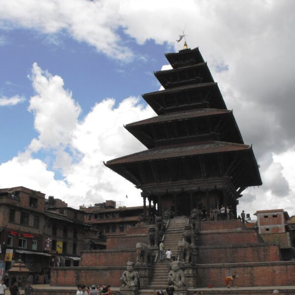 A multi-tiered temple dominates the foreground with a staircase leading up to it, flanked by stone statues, under a cloudy sky. Surrounding the temple are old traditional buildings with signs and people milling about in a city square.