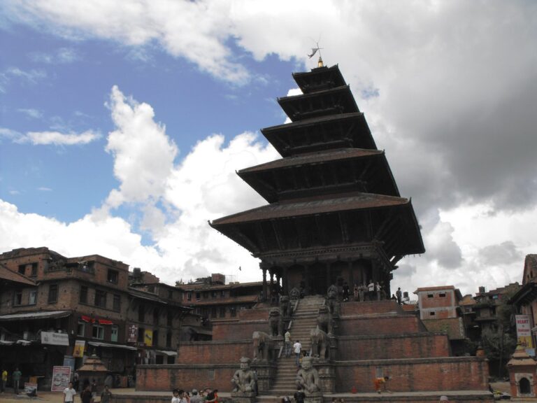 A multi-tiered temple dominates the foreground with a staircase leading up to it, flanked by stone statues, under a cloudy sky. Surrounding the temple are old traditional buildings with signs and people milling about in a city square.