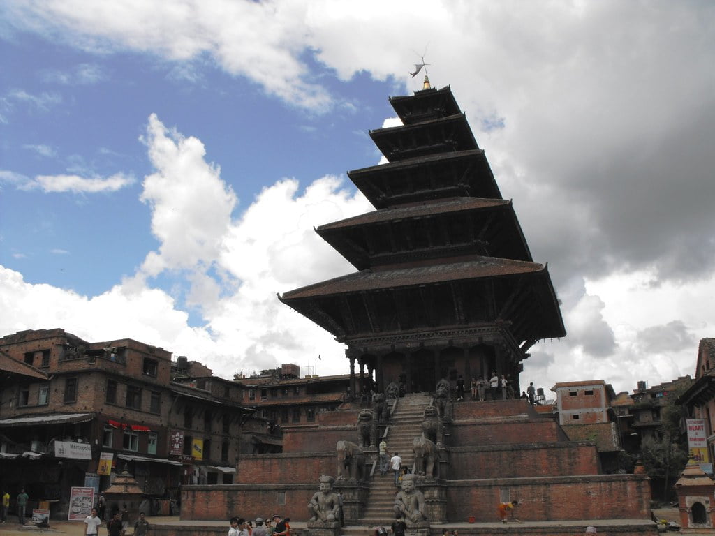 A multi-tiered temple dominates the foreground with a staircase leading up to it, flanked by stone statues, under a cloudy sky. Surrounding the temple are old traditional buildings with signs and people milling about in a city square.